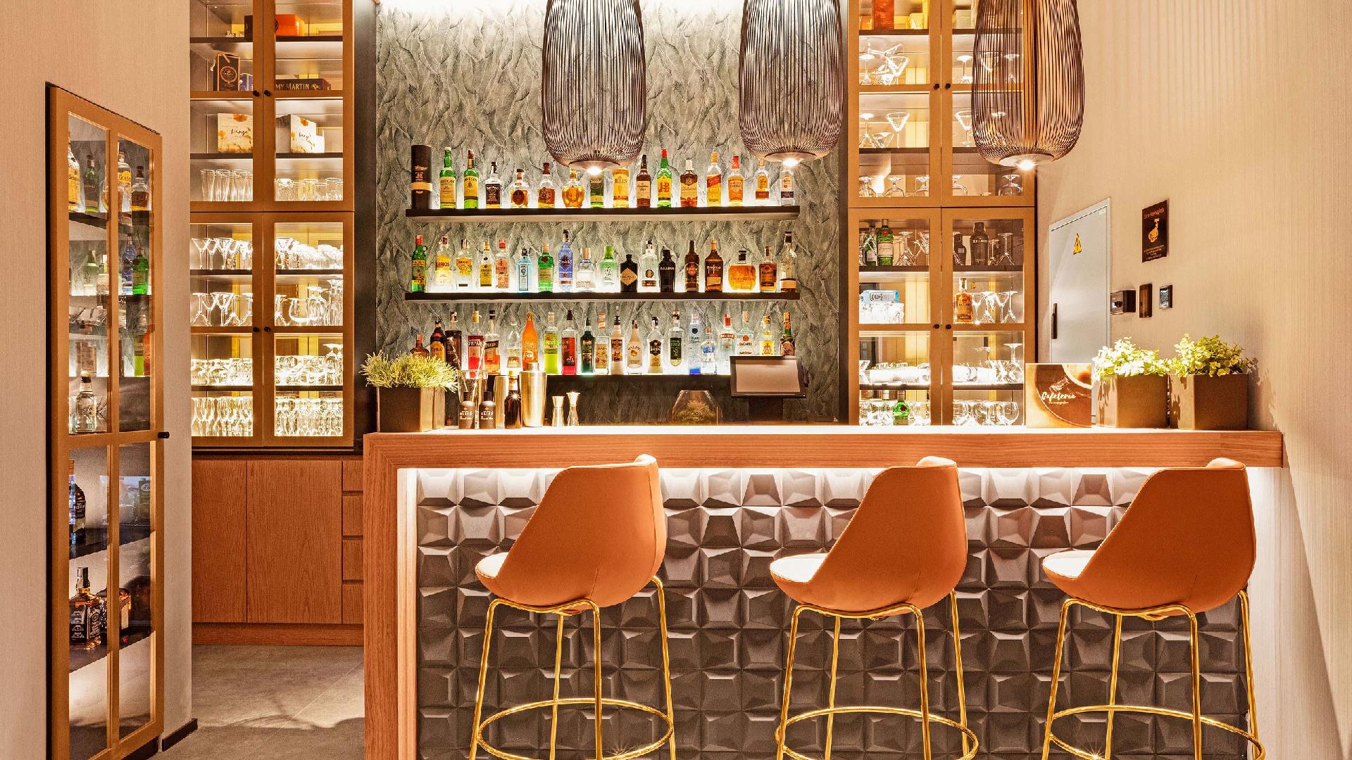Comptoir du café avec trois tabourets orange, étagères éclairées avec bouteilles et une enseigne qui indique Cafetería