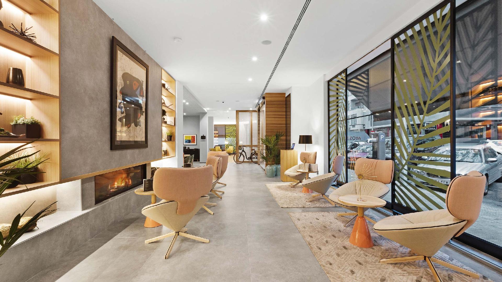Vestíbulo del hotel con chimenea encendida, un cuadro grande, sillones color melocotón y un ventanal con celosía de palmera