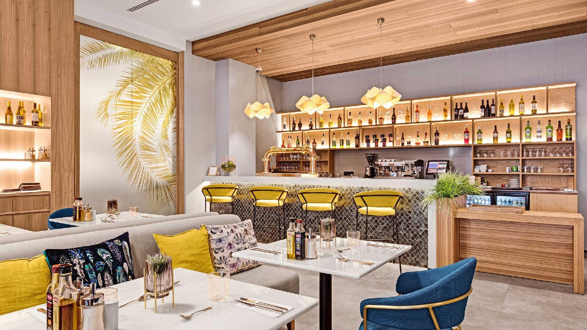 Hotel café with a bar with yellow stools, a golden palm mural, a grey sofa with cushions and blue armchairs