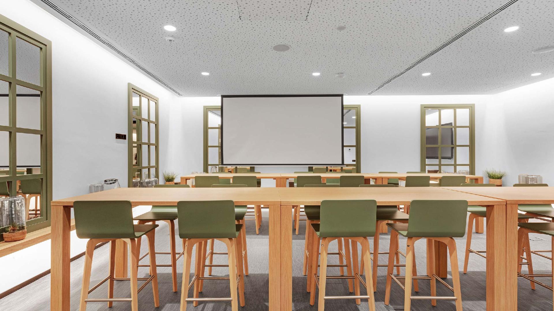 Sala con mesas altas y taburetes verdes, y una pantalla de proyección